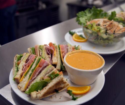 A plate of club sandwiches with lettuce, tomato, and ham, served with a bowl of orange soup and a side salad.
