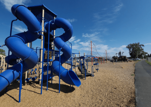 A colorful playground featuring blue slides, climbing structures, and a sandy area under a clear blue sky.