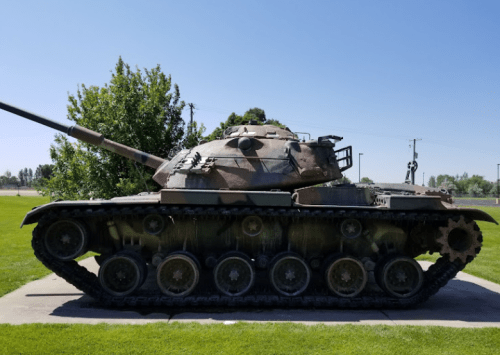 A side view of a weathered tank displayed on a grassy area, with trees and power lines in the background.