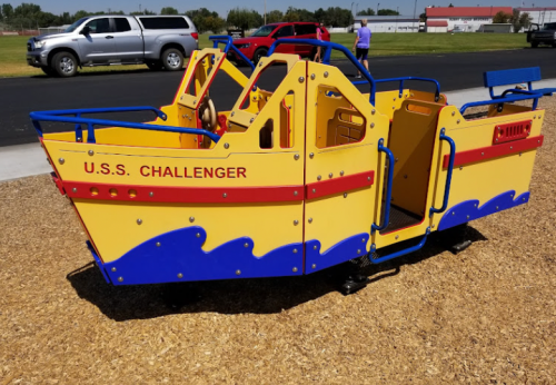 Colorful playground ship structure named "U.S.S. Challenger" with blue and yellow design, set on a sandy surface.