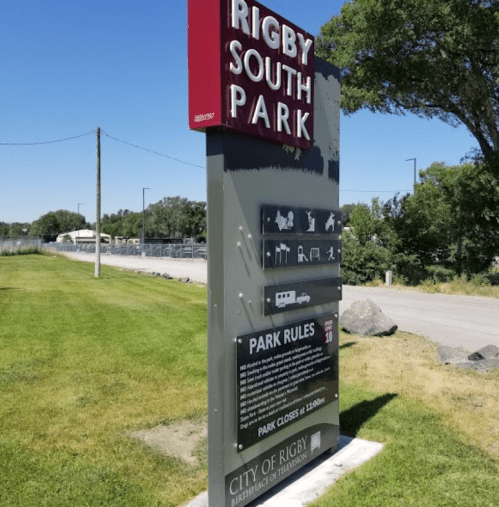 Sign at Rigby South Park displaying park rules and closing time, surrounded by green grass and trees.