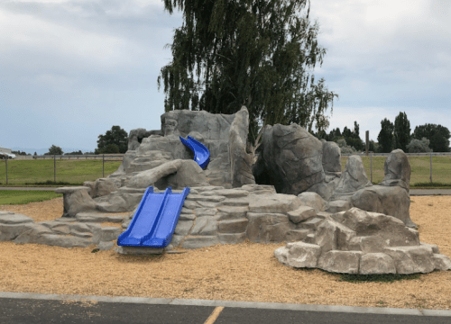 Playground structure resembling rocks with two blue slides, surrounded by gravel and grass under a cloudy sky.