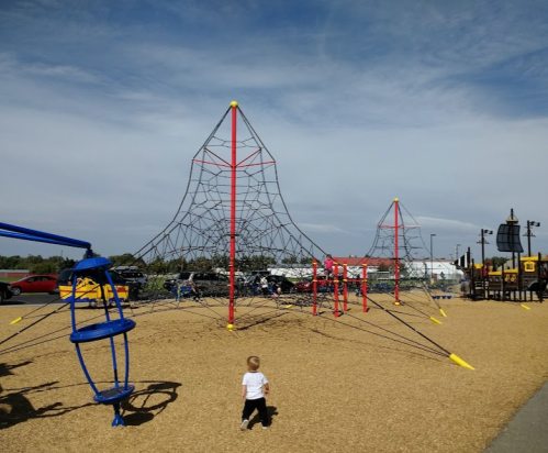 A child walks towards a large climbing structure in a playground, with blue and red equipment and a clear sky.