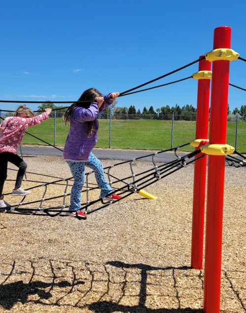 Two children play on a climbing structure at a playground, with a clear blue sky and grassy area in the background.