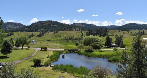 A scenic landscape featuring rolling green hills, a pond, and distant mountains under a clear blue sky.