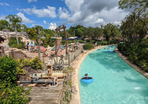 A lazy river at a water park, with people floating on tubes surrounded by lush greenery and sunny skies.
