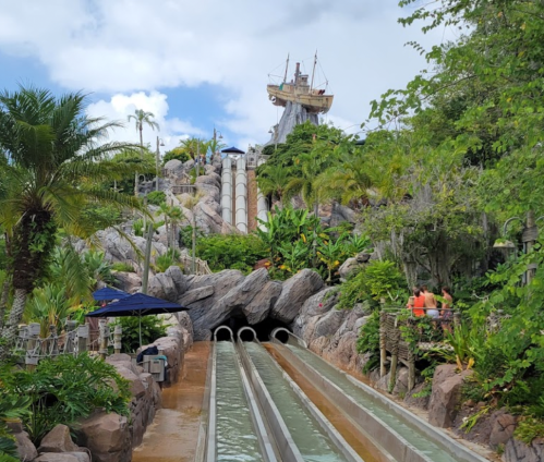Water slides surrounded by lush greenery and rocks, with a pirate ship structure in the background under a cloudy sky.
