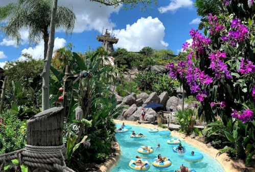 A tropical water park scene with people floating on inner tubes in a lazy river, surrounded by lush greenery and flowers.