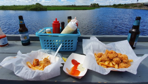A picnic setup by a river featuring fried chicken, fries, condiments, and drinks in a basket.