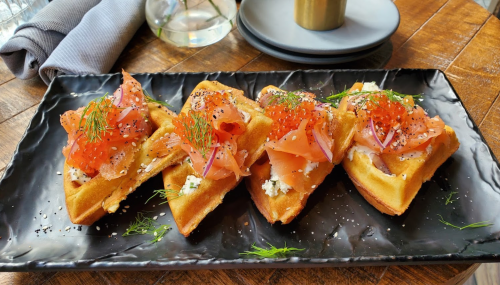 A black plate with four waffles topped with smoked salmon, cream cheese, red onion, and dill, garnished with roe.