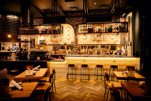 A stylish restaurant interior featuring a well-lit bar, wooden tables, and modern decor.