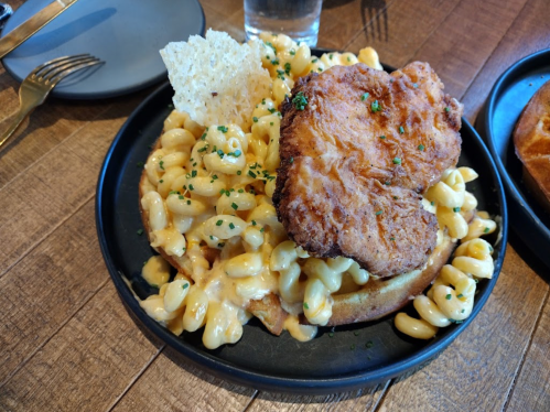 A plate of macaroni and cheese topped with fried chicken and a crispy garnish, served on a wooden table.