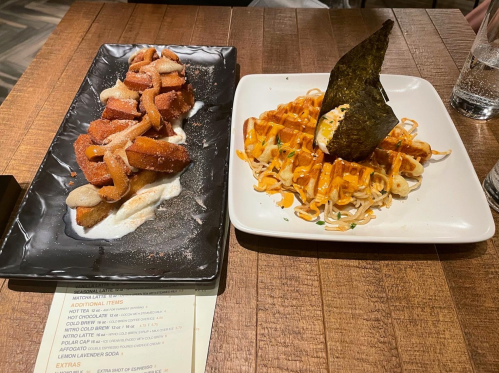 A plate with churros dusted in sugar and a plate of waffles topped with sauce and a seaweed garnish.