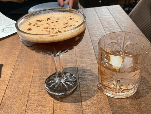 A cocktail in a coupe glass and a whiskey drink with a lemon slice on a wooden table.