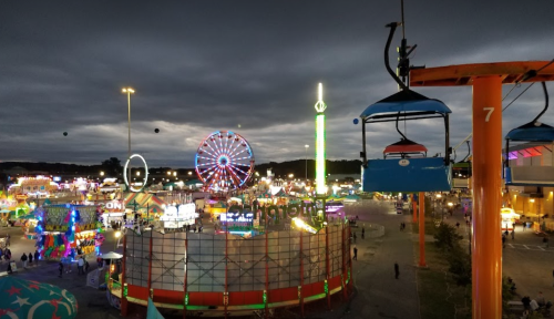 A vibrant fairground at night, featuring a Ferris wheel, colorful lights, and a cable car overhead against a cloudy sky.