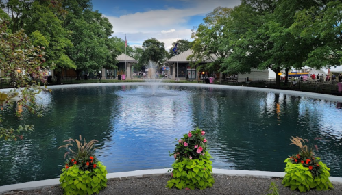 A serene pond surrounded by greenery and colorful flower planters, with buildings in the background under a cloudy sky.