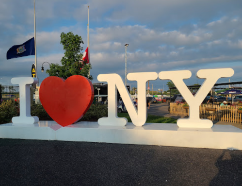 A large "I ❤️ NY" sign with a red heart, set against a cloudy sky and flags in the background.