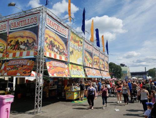 A bustling fair with colorful food stalls and banners, filled with people enjoying the festivities under a blue sky.