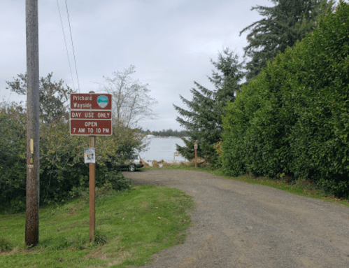 A gravel path leads to a sign for Prichard Wayside, indicating day use hours by a lake surrounded by trees.