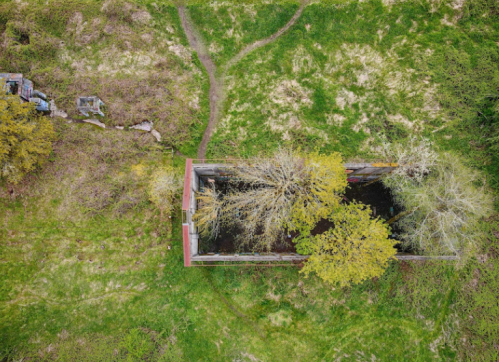 Aerial view of an abandoned structure surrounded by overgrown grass and trees, with a visible path nearby.