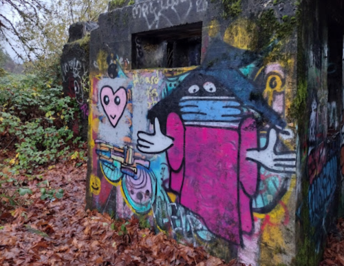 Colorful graffiti on a concrete wall featuring a character with a hat, mask, and heart, surrounded by foliage and fallen leaves.
