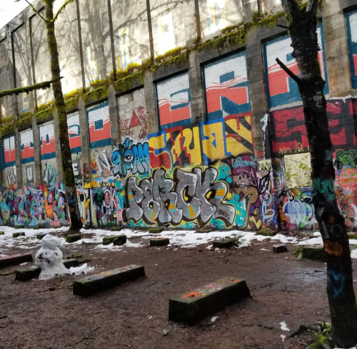 Colorful graffiti covers a wall in a wooded area, with patches of snow on the ground and trees nearby.