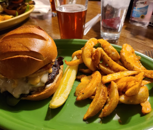 A burger with cheese and toppings on a green plate, accompanied by seasoned curly fries and a glass of beer.