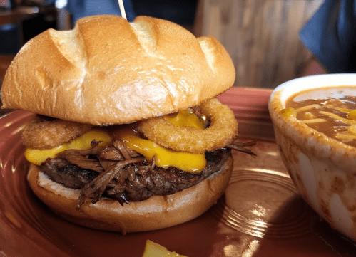 A close-up of a burger with a beef patty, melted cheese, and onion rings, served with a bowl of chili on the side.
