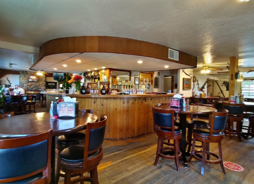 Cozy restaurant interior with wooden bar, tables, and decor, featuring a well-stocked bar and a warm atmosphere.