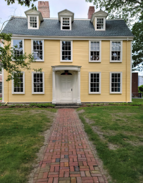 A yellow two-story house with white trim, large windows, and a brick pathway leading to the front door.