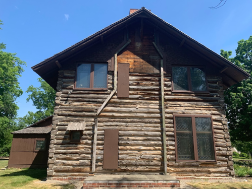 A rustic log cabin with multiple windows and doors, surrounded by greenery under a clear blue sky.