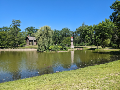 A serene park scene featuring a pond, a lighthouse, lush trees, and a clear blue sky.