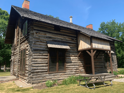 A rustic log cabin with a sloped roof, large windows, and a picnic table in front, surrounded by greenery.