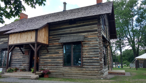A rustic log cabin with a wooden balcony, surrounded by greenery and flowers, under a partly cloudy sky.