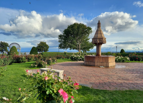 A scenic park featuring a decorative fountain surrounded by lush greenery and blooming roses under a blue sky.