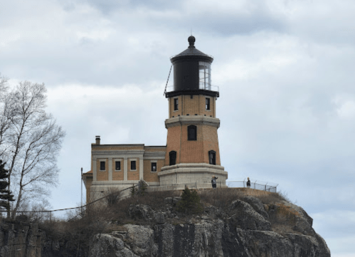 A historic lighthouse atop a rocky cliff, surrounded by trees and cloudy skies.