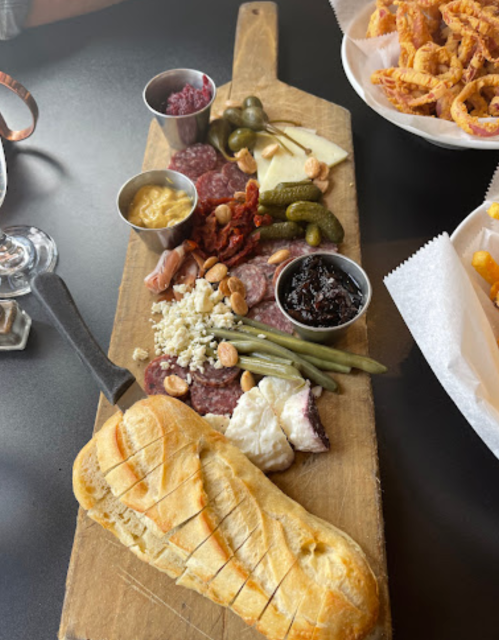 A wooden board with assorted meats, cheeses, pickles, nuts, and bread, accompanied by dipping sauces.