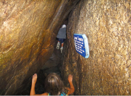 A narrow rock crevice with a child climbing through, and a sign on the wall providing information about the location.