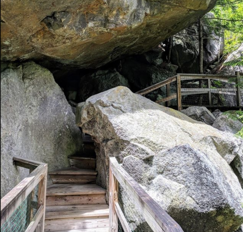 A wooden walkway leads through large boulders and rocks in a natural setting, surrounded by greenery.