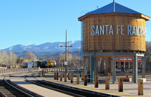 A wooden water tower labeled "Santa Fe Railway" beside train tracks, with mountains in the background on a clear day.