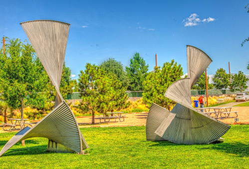 Two large, abstract metal sculptures in a grassy park, surrounded by trees and picnic tables under a clear blue sky.