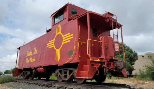 A red caboose with a yellow New Mexico symbol, parked on tracks under a cloudy sky.