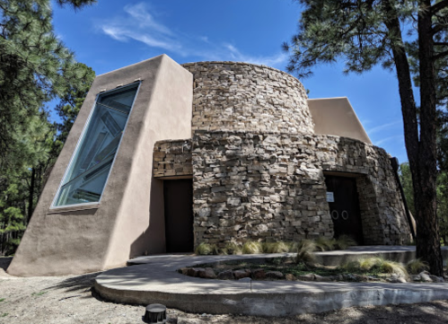 A modern stone and adobe building surrounded by trees, featuring a large angled window and a circular design.