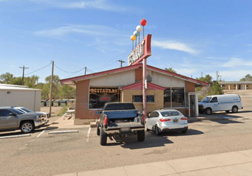 A small restaurant with a red roof and colorful balloons, surrounded by parked cars on a sunny day.