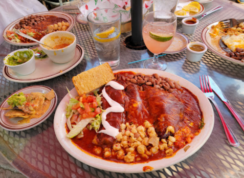 A colorful plate of enchiladas with beans, corn, and salad, accompanied by drinks and a side of guacamole.