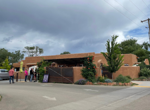 A southwestern-style building with adobe walls, surrounded by greenery, and people gathered outside.