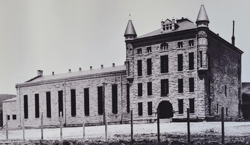 Historic stone building with towers, large windows, and a fenced perimeter, set against a clear sky.
