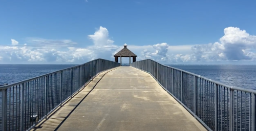 A long pier extends over calm water, leading to a small gazebo under a blue sky with fluffy clouds.