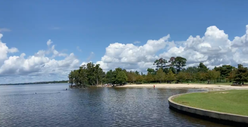 A serene lakeside view with trees, sandy beach, and fluffy clouds in a blue sky.