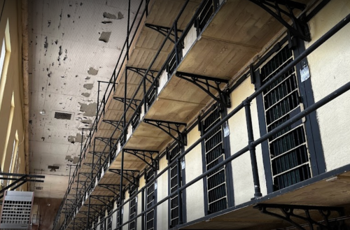 Interior view of a prison cell block with multiple cells and peeling paint on the ceiling.